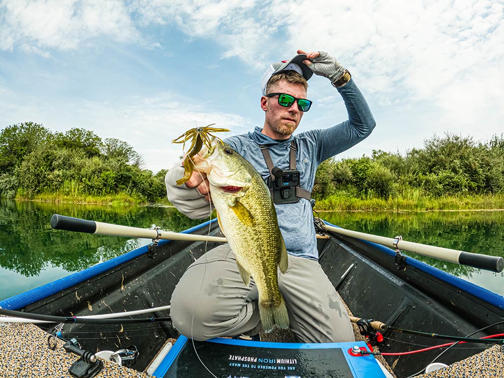 A man sitting in a small boat on a lake holding up a big Smallmouth Bass he caught, with fishing gear and oars around him.