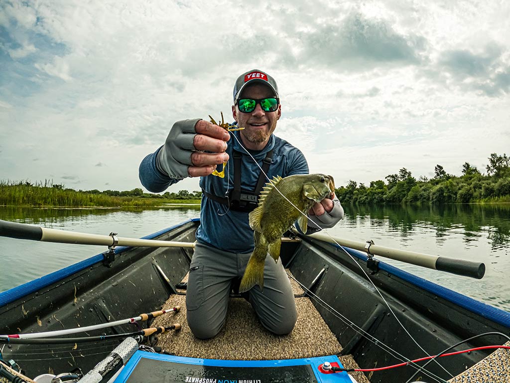 A man kneeling in a boat, holding a small Bass in one hand and fishing gear in the other, with water and lush greenery behind them, showing off fishing with a free rig