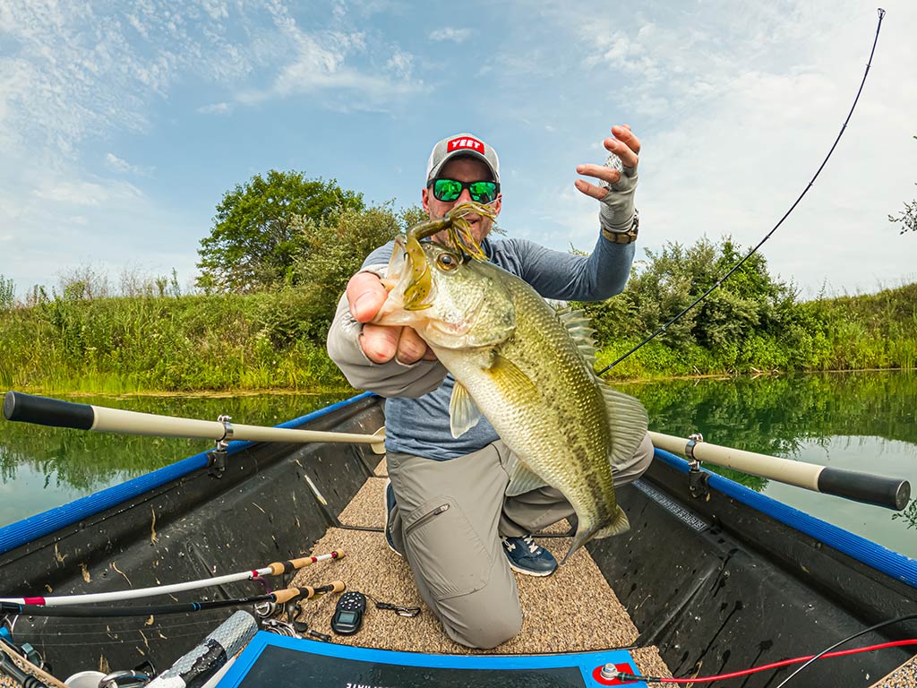 A man kneeling in a small boat, holding a Bass with a fishing rod in hand, while water and greenery are in the background.