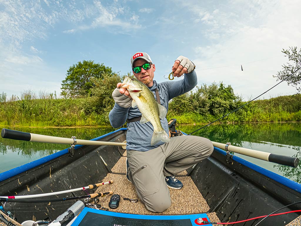 A man kneeling in a boat on a lake, holding a Bass in one hand and a fishing lure in the other, with fishing gear close by.