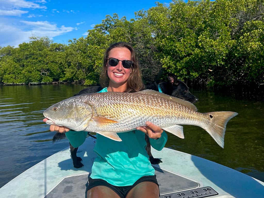 A woman enjoying summer fishing in the Everglades, smiling at the camera as she holds a large Redfish on a boat with trees and water in the background.