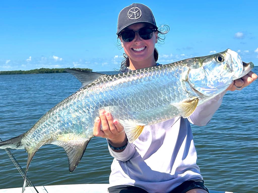 A woman wearing sunglasses and a hat proudly holds a large silver Tarpon on a boat under a clear blue sky.