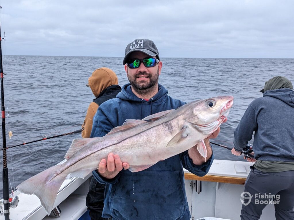 A man’s holding a large Haddock on a boat during New Hampshire's fishing deason, while two others are casting their lines in the background on a cloudy day.