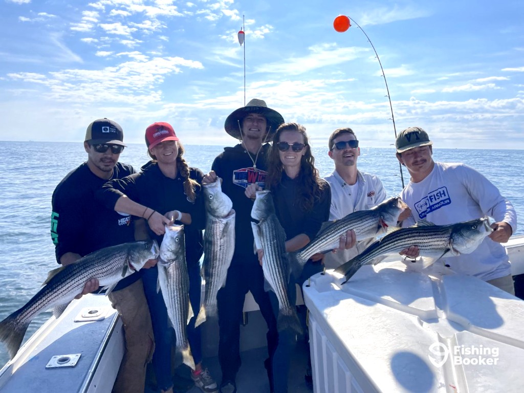 Six people stand in a row on a boat, each holding a large Striped Bass, with big smiles and arms proudly extended to display their catches. The sea stretches out behind them under a partly cloudy sky, creating a lively backdrop for their celebration.