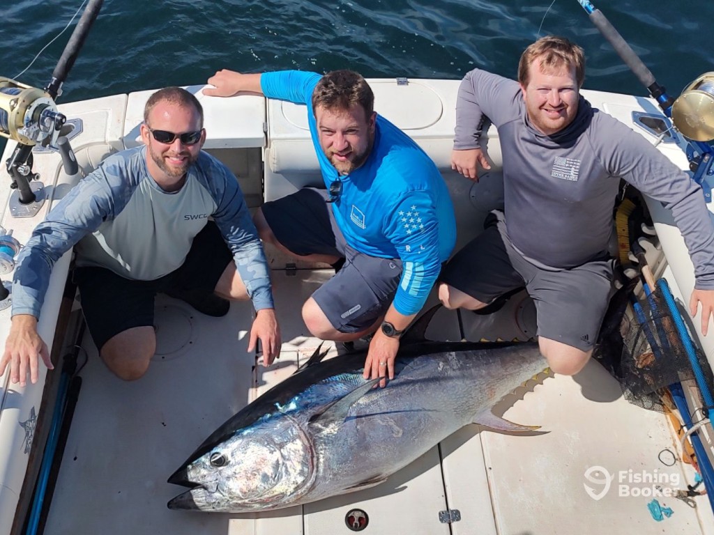 Three men on a fishing boat pose with a large Tuna they've caught during New Hampshire's offshore fishing season. Fishing rods are visible, and the sea stretches out in the background.
