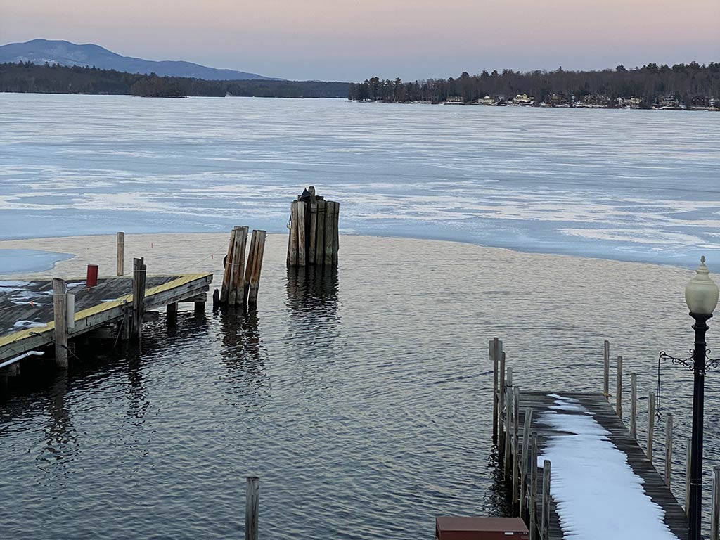 A partially frozen lake stretches out in New Hampshire, its wooden docks and pilings jutting above the icy surface. Some of the ice is melting near the shore, creating open water.