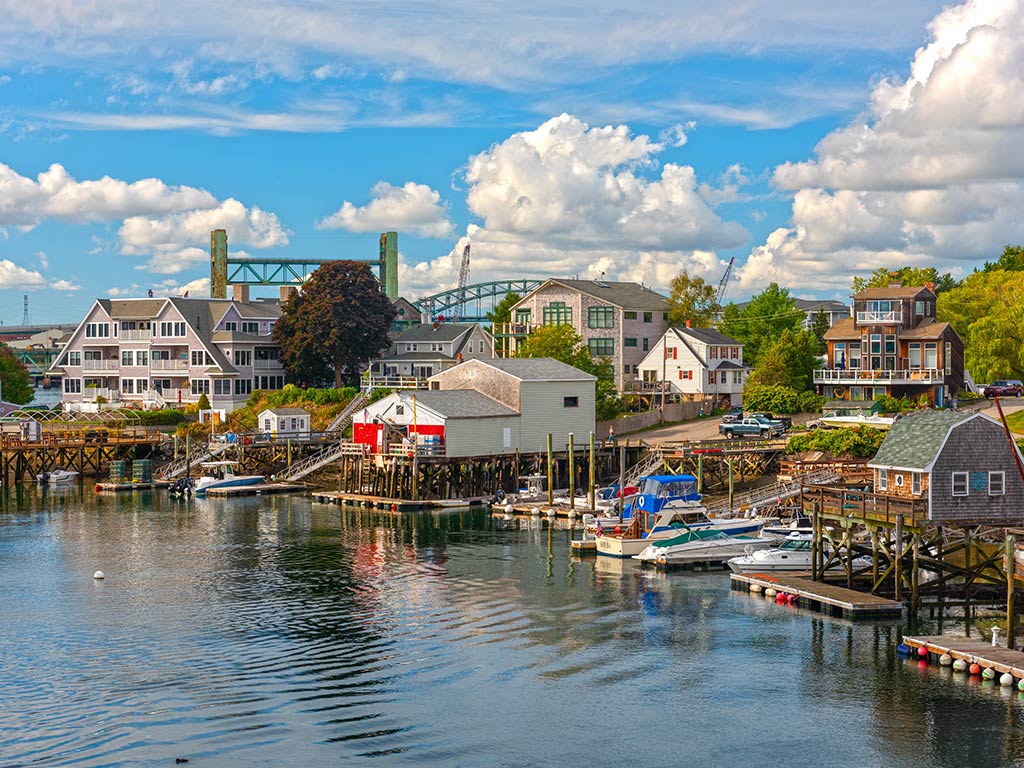A small harbor in Portsmouth, NH features docked boats, wooden piers, and waterfront houses under a partly cloudy sky. In the background, a green bridge rises above the scene—its reflection shimmering on the water.