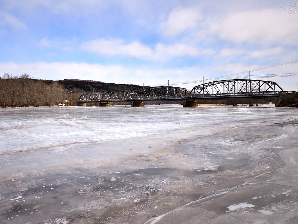 A steel truss bridge spans across a frozen river under a partly cloudy sky, with bare trees and hills in the background.