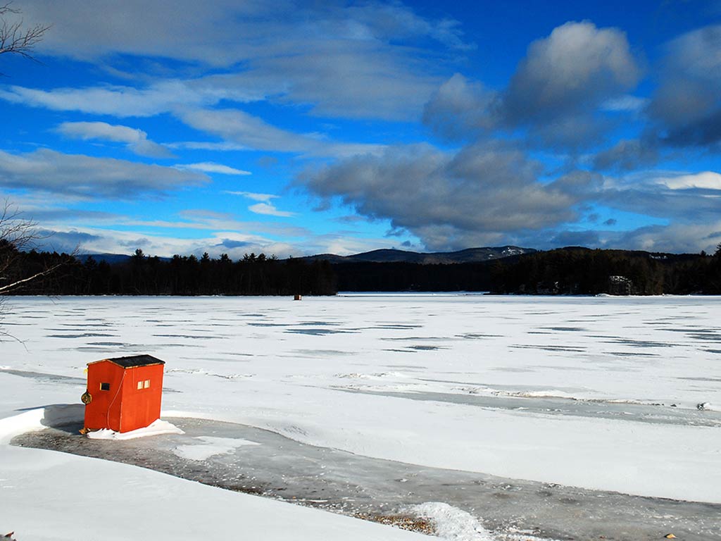 A small red ice fishing hut sits on a frozen, snow-covered lake under a partly cloudy sky.