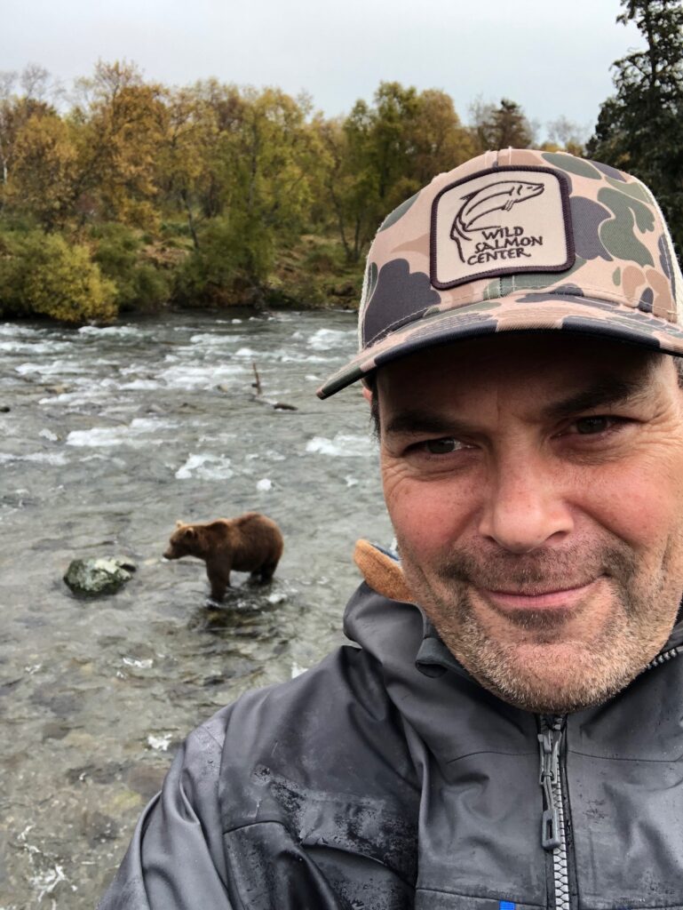 A man in outdoor clothing's taking a selfie by a river, with a Brown Bear standing in the water behind him.