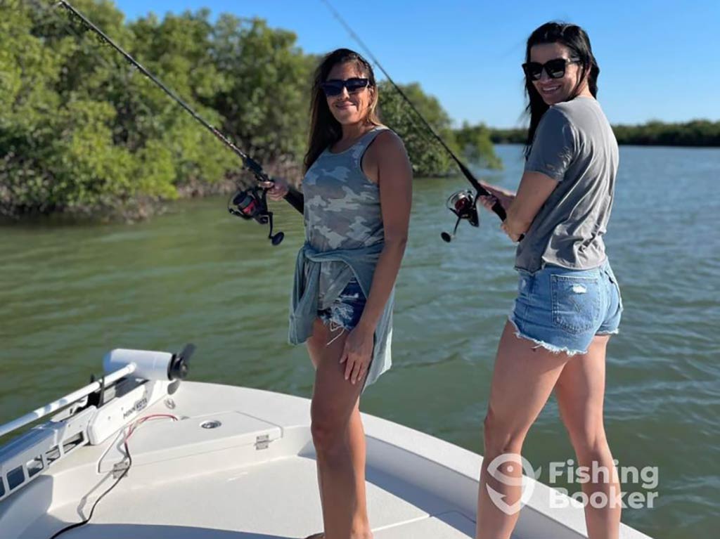 Two women are on a boat holding fishing rods, facing the water, with greenery and trees behind them on a sunny day.