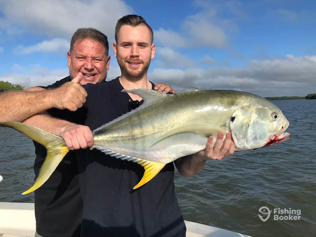 Two men on a boat hold a large Jack Crevalle., as one man is giving a thumbs up. Water and trees are in the background on a clear day.