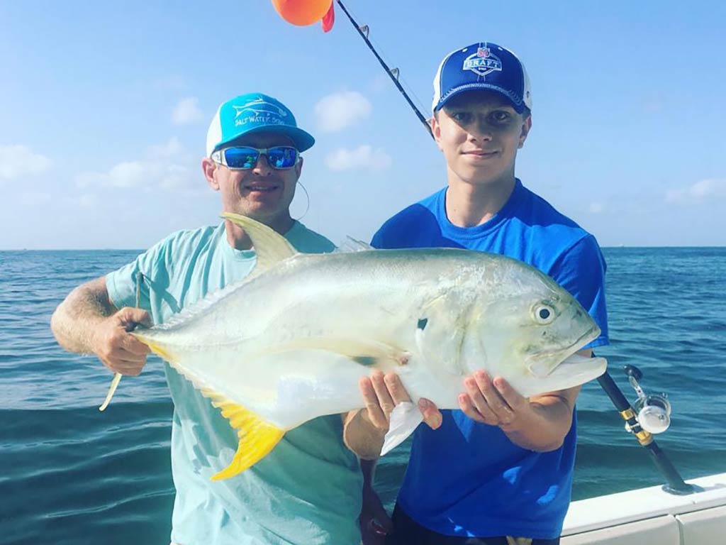 Two people on a boat are holding a large Jack Crevalle fish, with the ocean and blue sky in the background.