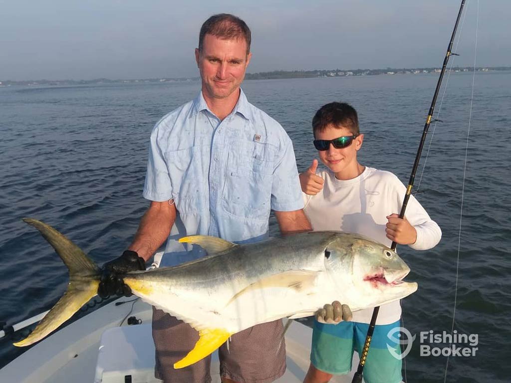 A man and a boy on a boat holding a large Jack Crevalle. The adult has the fish while the child is holding a fishing rod and giving a thumbs up. Water and distant shoreline are in the background.