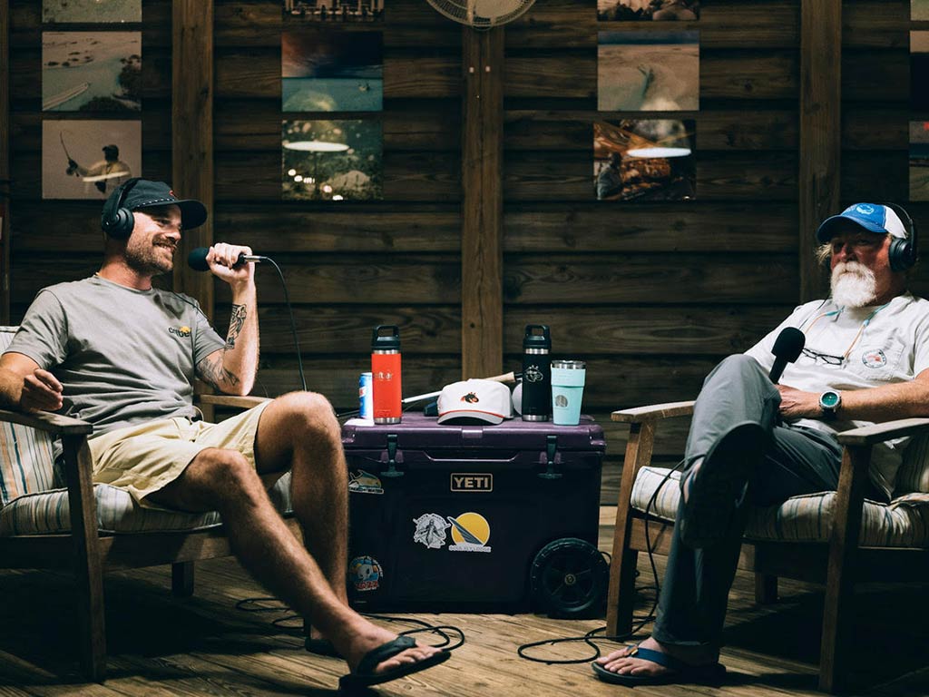 Two men sit in chairs with microphones and headphones, recording one of the best fishing podcasts in a wooden room with photos on the wall and a YETI cooler between them.
