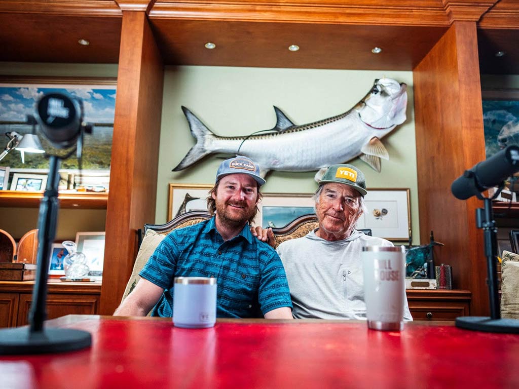 Two men wearing caps sit together in front of a large mounted fish, recording a fishing podcasts with microphones and drink tumblers on a red table in the foreground.