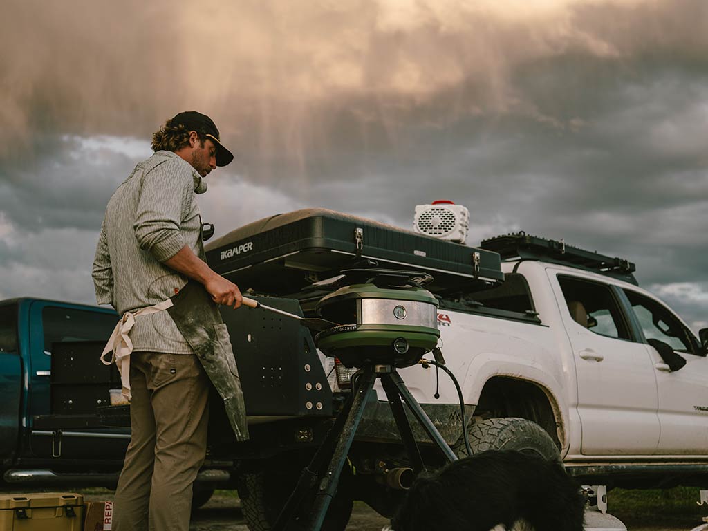 A man standing by a white pickup truck using a portable grill under overcast skies, while his black-and-white dog hangs out next to him.