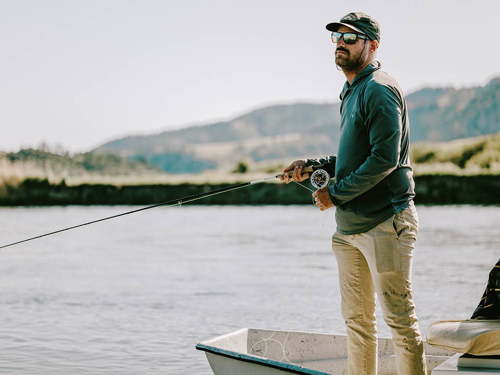 A man wearing sunglasses and a cap stands on a small boat holding a fishing rod with a river and hills in the background.