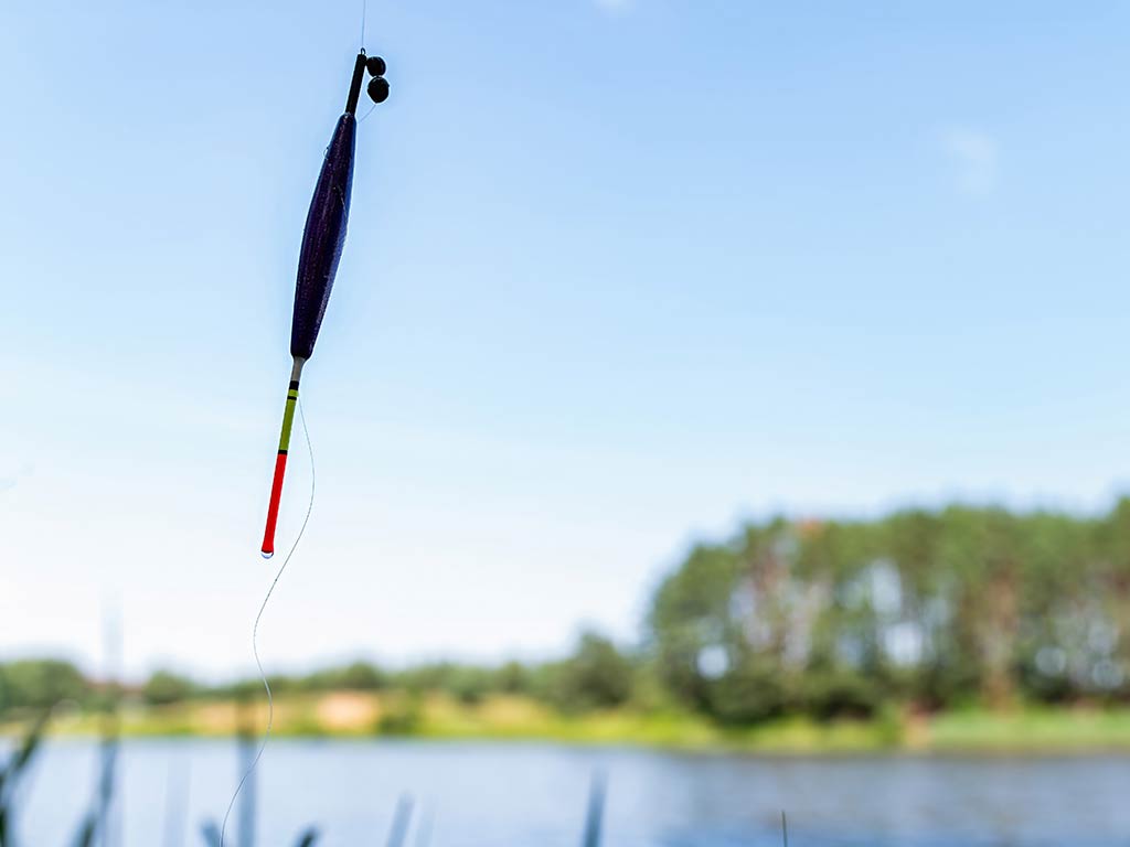 A fishing bobber float hanging on a fishing line in the foreground, while a blurred lake and trees fill the background.