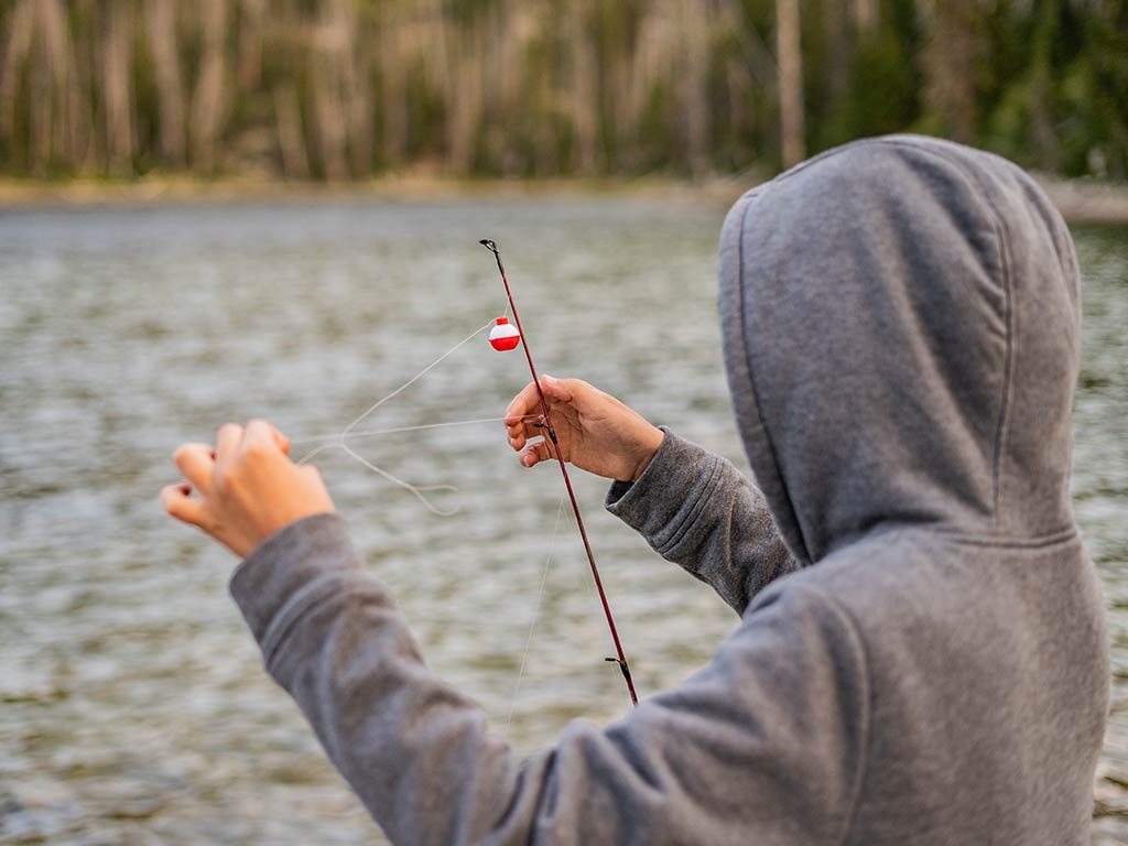A view from behind of someone in a gray hoodie standing by a body of water, bobber fishing with a red and white bobber, and they're adjusting the fishing line on their rod.