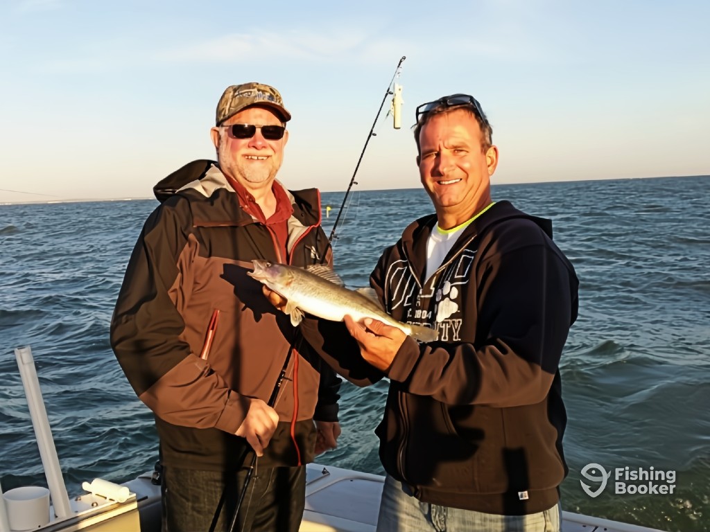 Two men on a boat are smiling at the camera; one's got a fishing rod and bobber, while the other shows off a freshly caught Speckled Trout. The ocean and sky can be seen in the background.