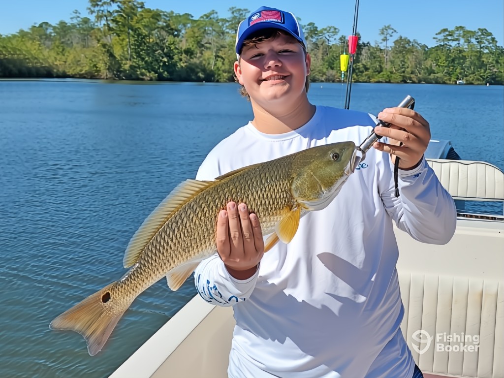 A man on a boat, holding up a large Redfish with one hand and gripping a fish scale with the other.