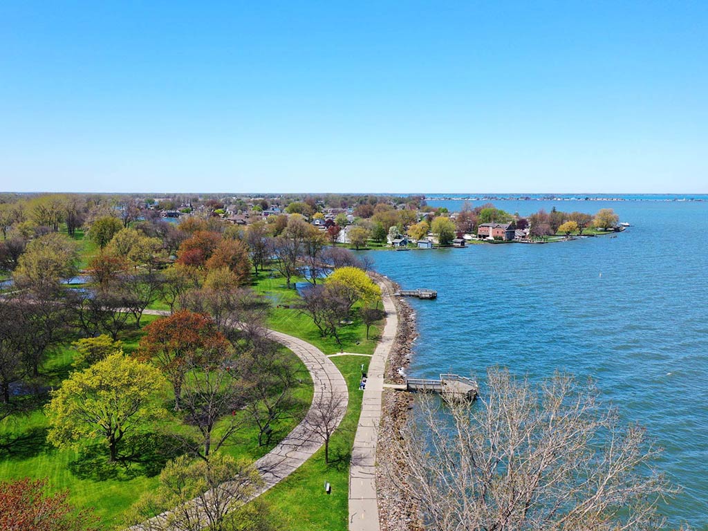 An aerial view of the Metropark alongside Lake St. Clair on a clear day, with winding walking paths, green trees, and waterfront homes visible.