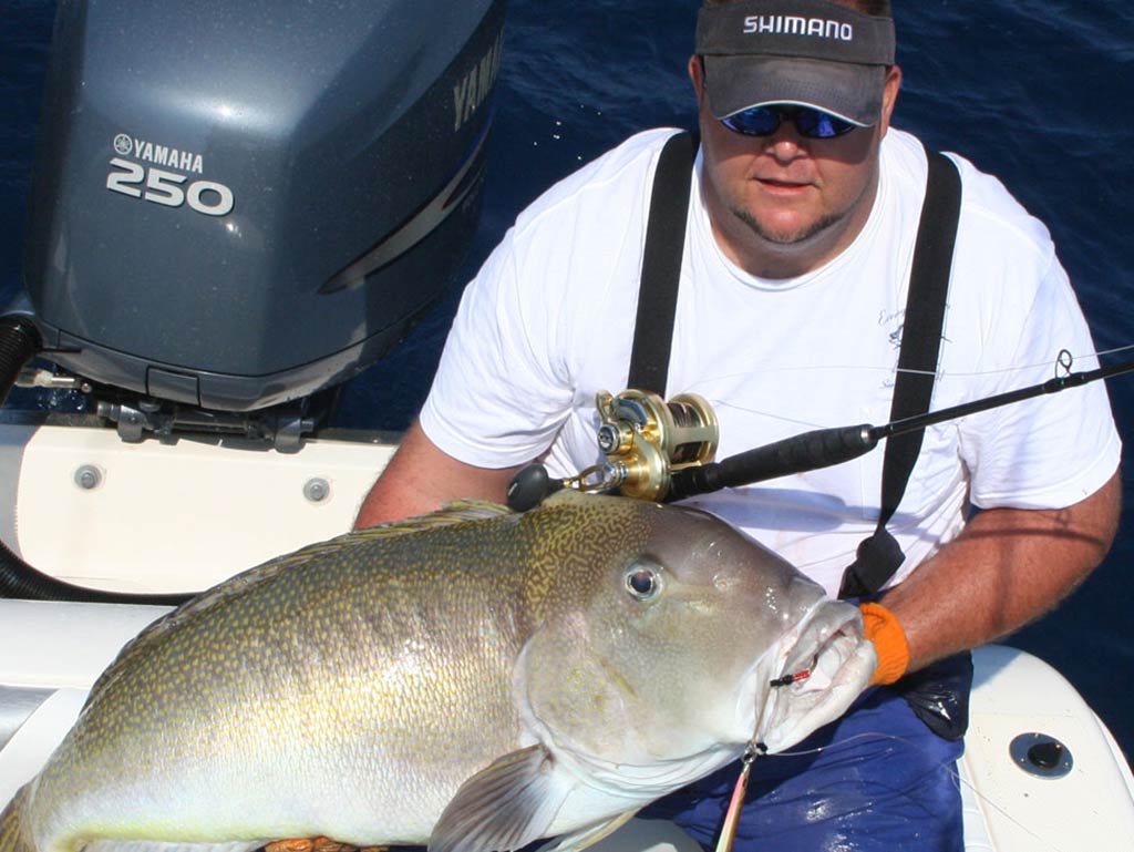 A man in a white shirt and cap holding a large Tilefish on his lap on a boat near an outboard motor, with fishing equipment visible.