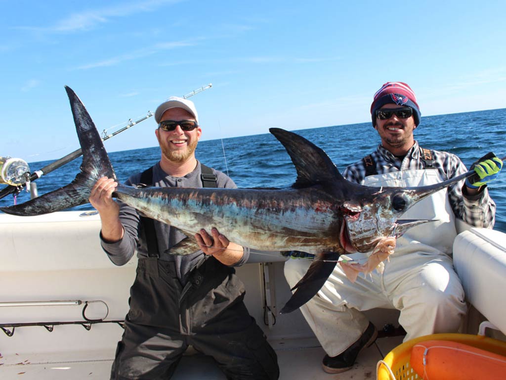 Two people on a boat are holding a large Swordfish and smiling at the camera. You can see fishing equipment and the ocean in the background on a sunny day.