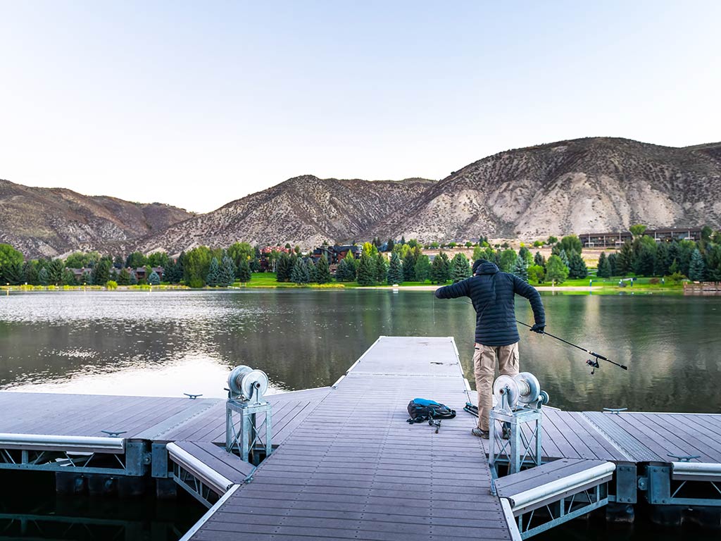A view from behind of someone in a jacket standing on a dock, casting a fishing line into the calm waters of Beaver Creek, while mountains and trees are in the background.