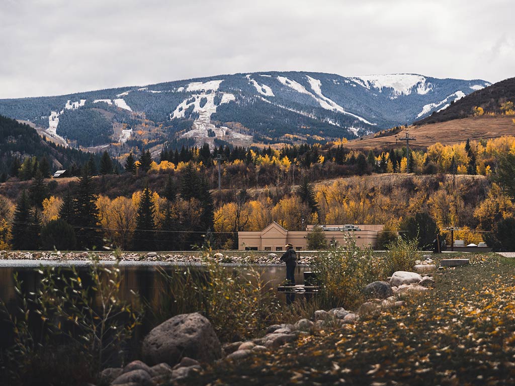 A view towards someone fishing by a lake at Beaver Creek, surrounded by fall-colored trees, a building, and snow-patched mountains under a cloudy sky.