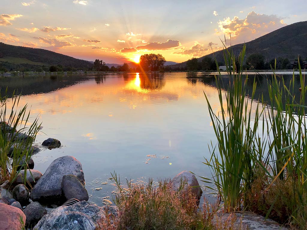 A view of a calm lake at sunset near Beaver Creek, CO, with the sun setting behind distant hills and shining on the water. Tall grass and rocks line the shore.