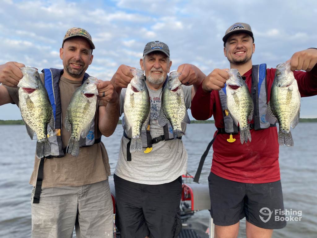 Three men wearing life jackets are holding up freshly caught Crappie while standing on a boat, with a lake and cloudy sky in the background.