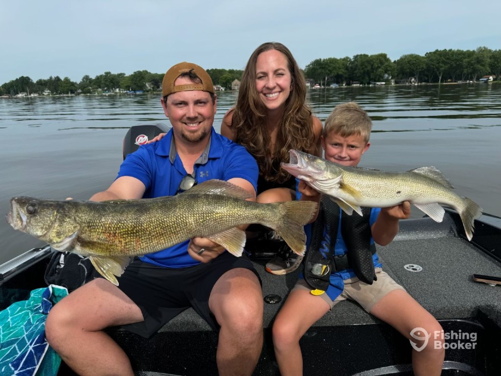 A family of three people on a boat are holding up two Walleyes they caught in Illinois. They're smiling at the camera, with a lake and trees in the background.