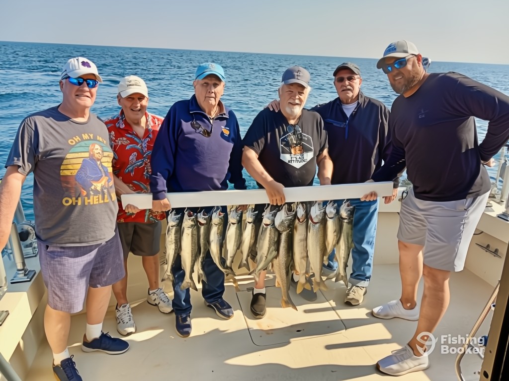 Six men on a boat show off a row of freshly caught Salmon, enjoying summer – one of the best Illinois fishing seasons –with a Great Lake visible in the background.