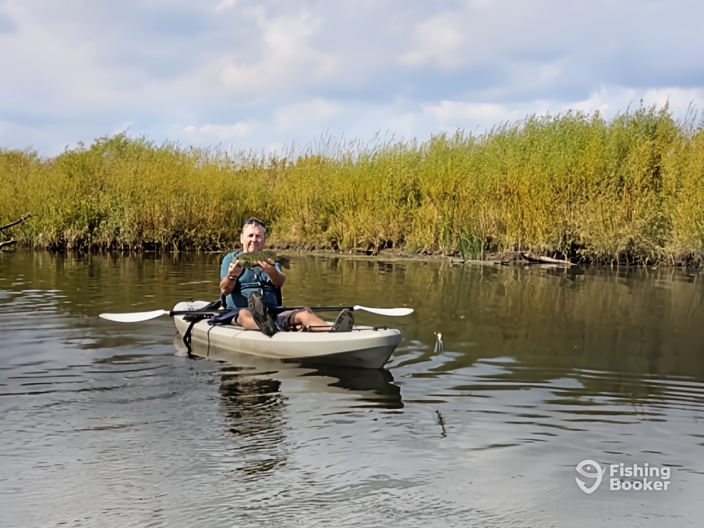 A man sitting in a kayak on calm water, holding up a Bass and enjoying the surroundings, with reeds and bushes visible behind him.