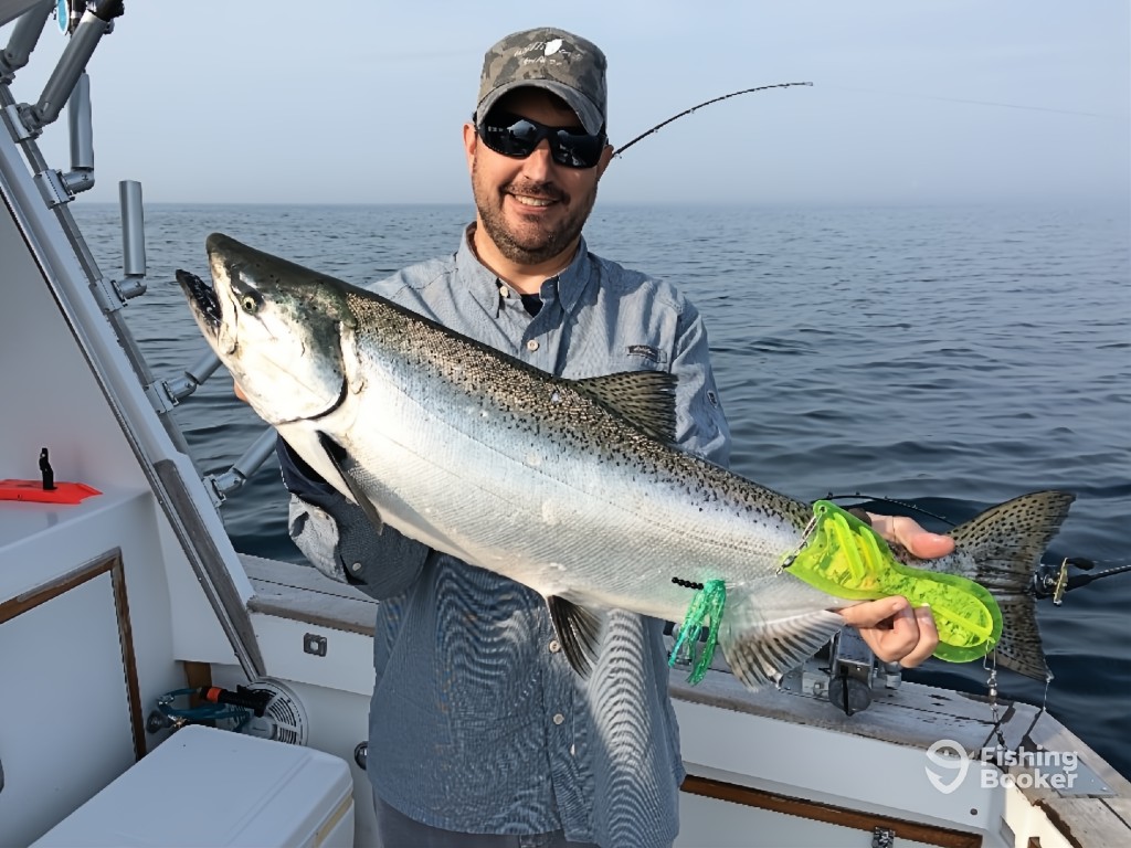 A man standing on a boat, holding a large Salmon with dark spots as he smiles at the camera. The vast expanse of one of the Great Lakes is visible in the background.