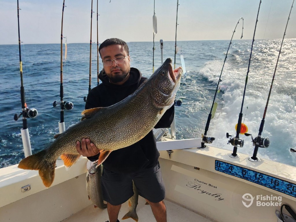 A man standing on a boat holding a large Lake Trout with several fishing rods and the open ocean are behind him.