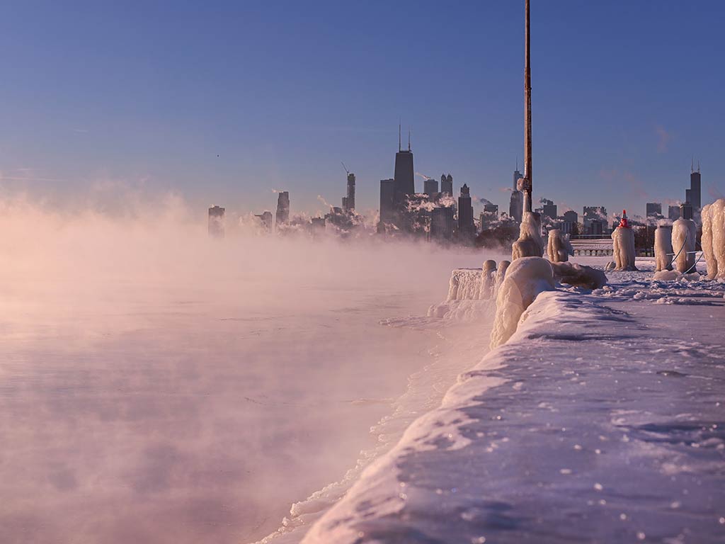 A frozen lakefront with snow and ice up front, mist rising off the water, and the skyline of Chicago visible under a clear sky.