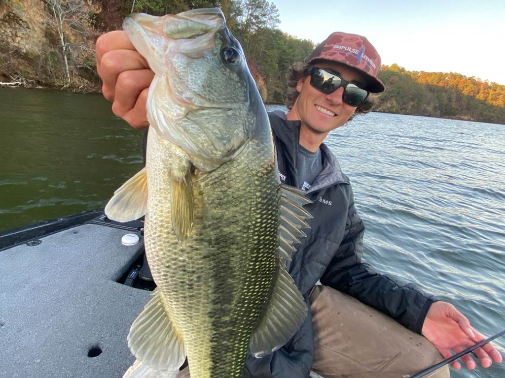 A man sitting in a boat on a lake, holding a large Bass with one hand and smiling at the camera. Trees and the water of Lake Guntersville, AL are in the background.