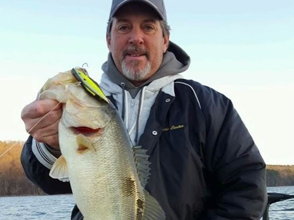 A man in a jacket and cap's holding a large Bass with a lure in its mouth, standing outdoors near Lake Guntersville.