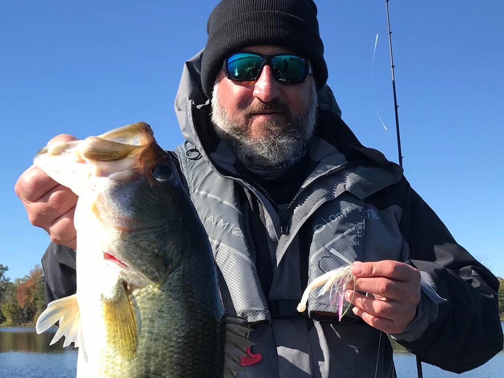 A man in outdoor gear holding a large Bass in one hand and a fishing lure in the other, standing by Lake Guntersville on a clear day.