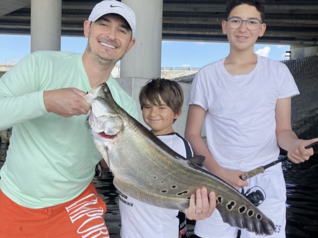 A man, teenager, and child standing on a dock while showing off their Clown Knife Fish catch.
