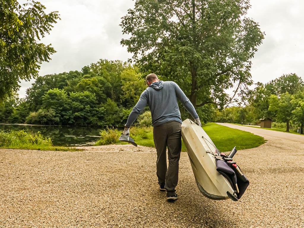 A view from behind of a man lugging a kayak across a gravel path towards a pond on a cloudy day.
