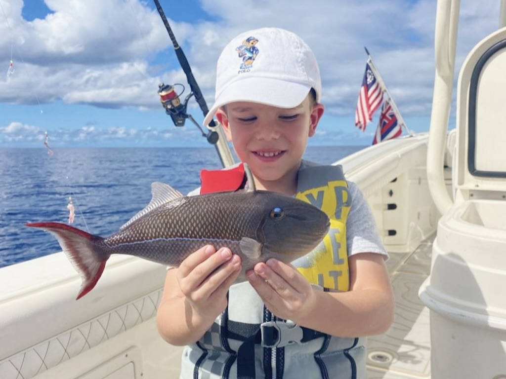 A child in a white baseball cap and yellow life jacket holds up a colorful Blueline Triggerfish as he stares at it intently, with flags and the ocean visible behind him.