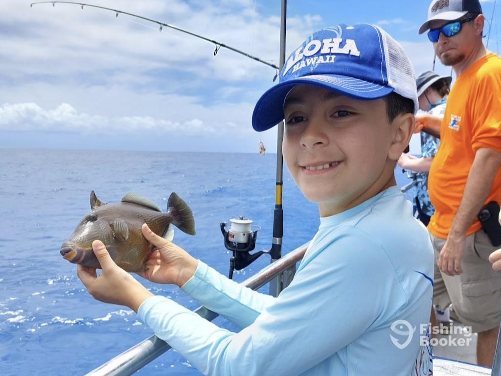 A child in a baseball cap holding up a small Triggerfish over the side of a fishing boat, as someone fishes behind them.