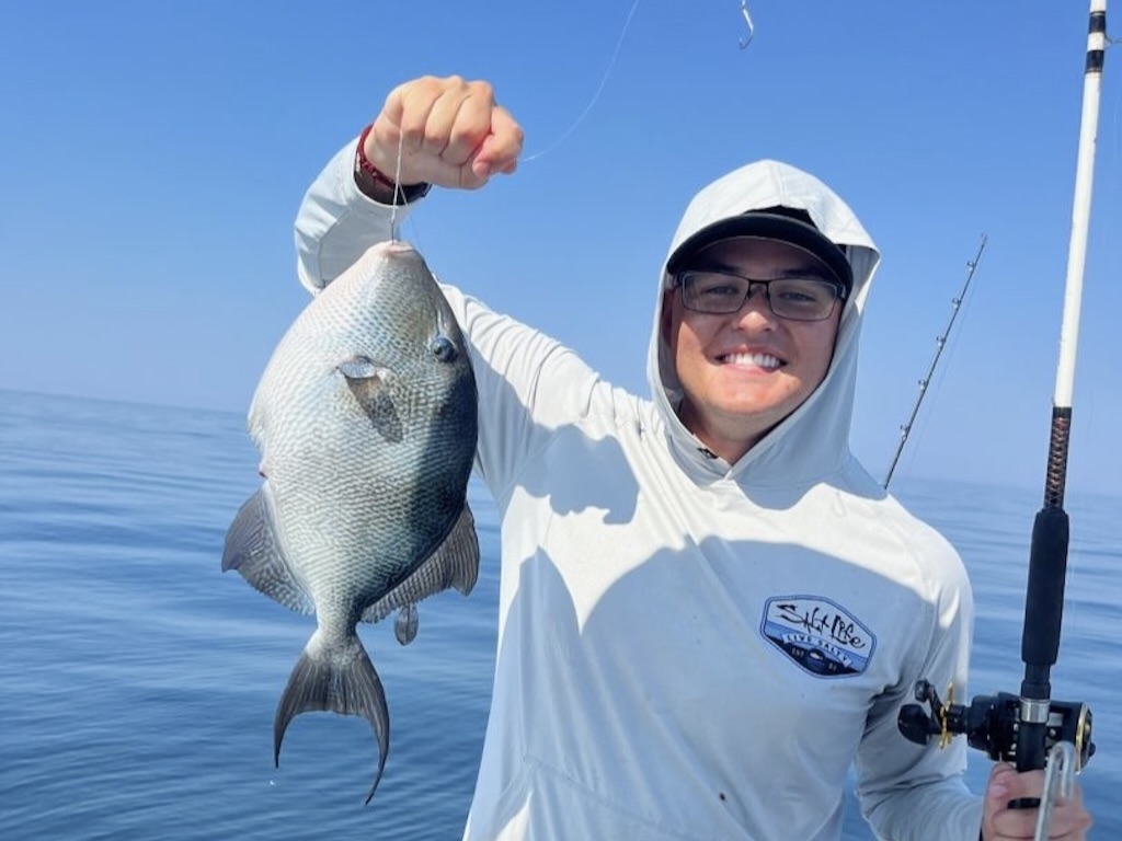 A young man in a hoodie and wearing glasses, holding up a Triggerfish by the line he caught it with in his right hand on a boat on a clear day.