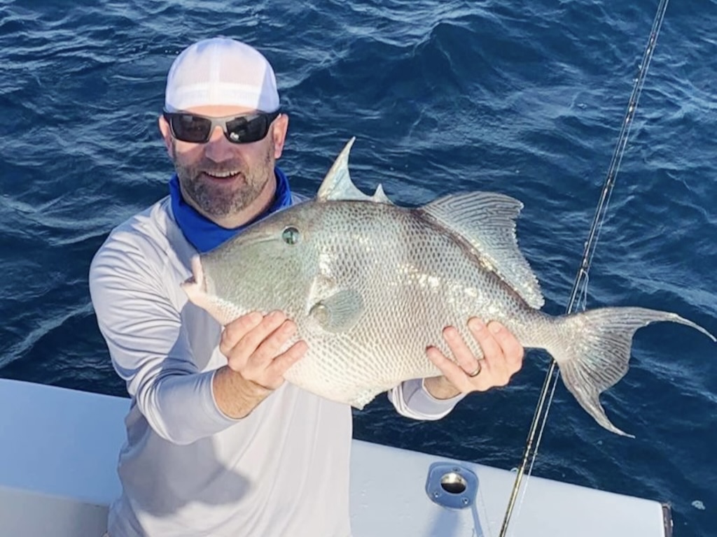 A man in a bandana and sunglasses holding up a sizeable Triggerfish to the camera while standing on a boat.