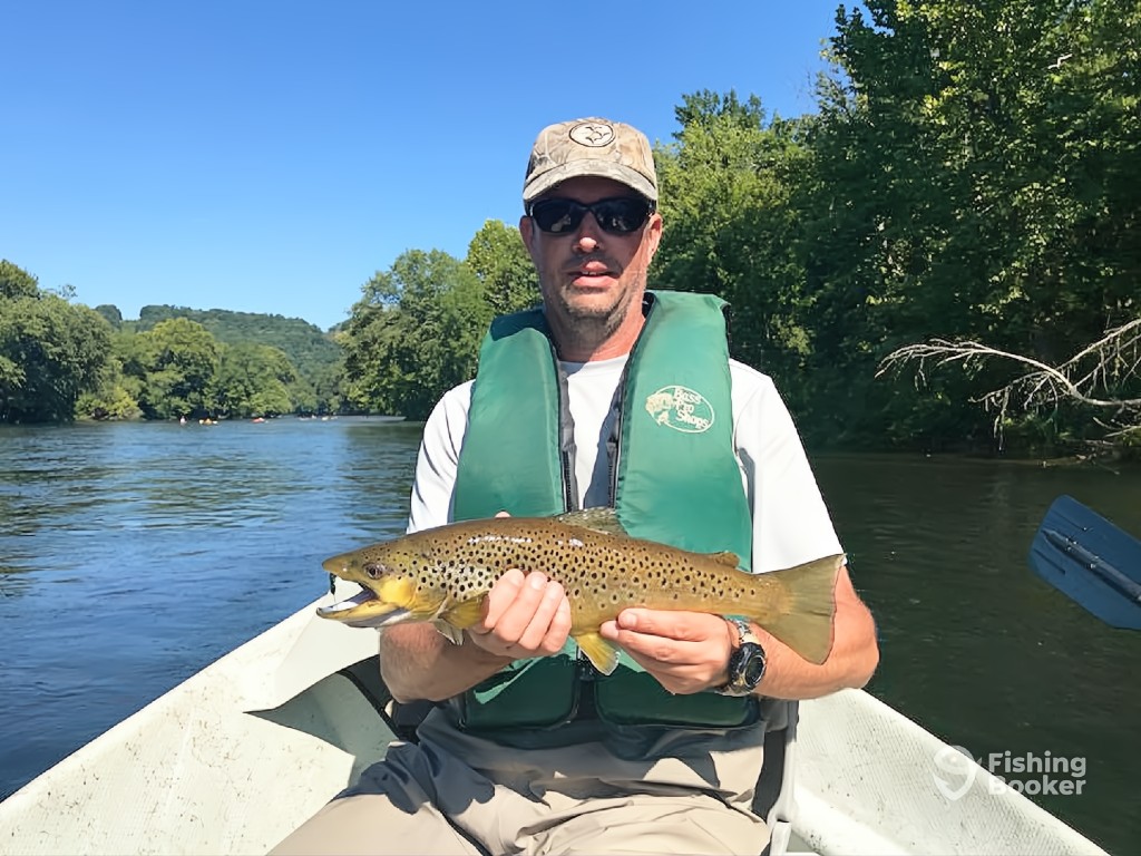 A man in sunglasses and a life vest's sitting in a boat on a river, holding a large Brown Trout—an iconic moment of Trout Fishing in Tennessee, with trees and hills behind him.