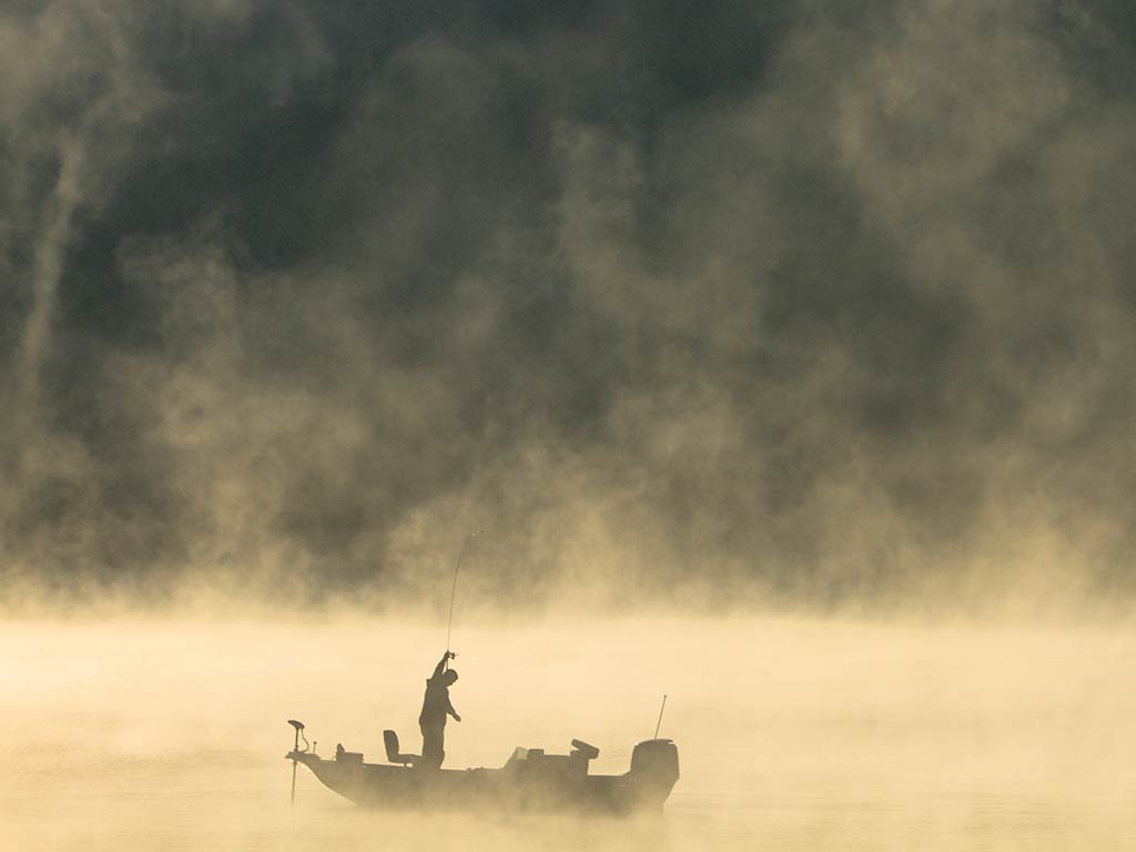 A view across a water at dawn, with a silhouette of an angler on a boat fishing in Tennessee on a river surrounded by thick morning mist drifting over the water.